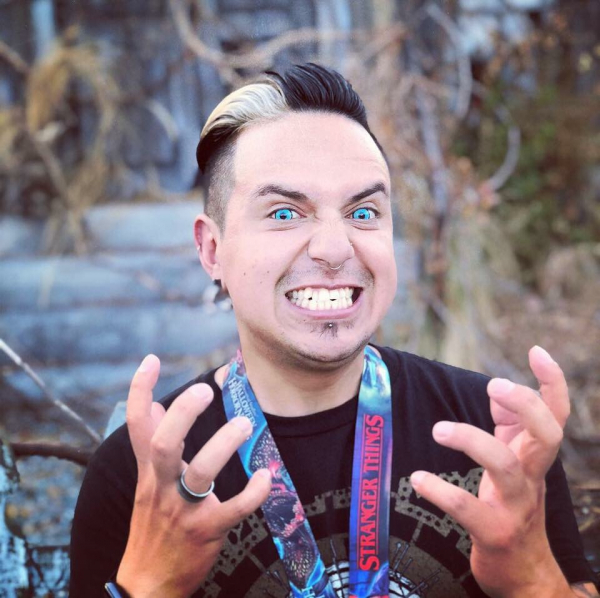 A photograph of a person wearing bright blue contacts bares their teeth at the camera. Both their hands are lifted up and making claw shapes. They have short dark hair with a white streak in it.