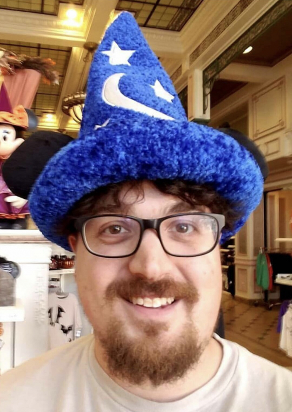 A smiling person with a blue wizard hat on. The person has a beard and glasses.