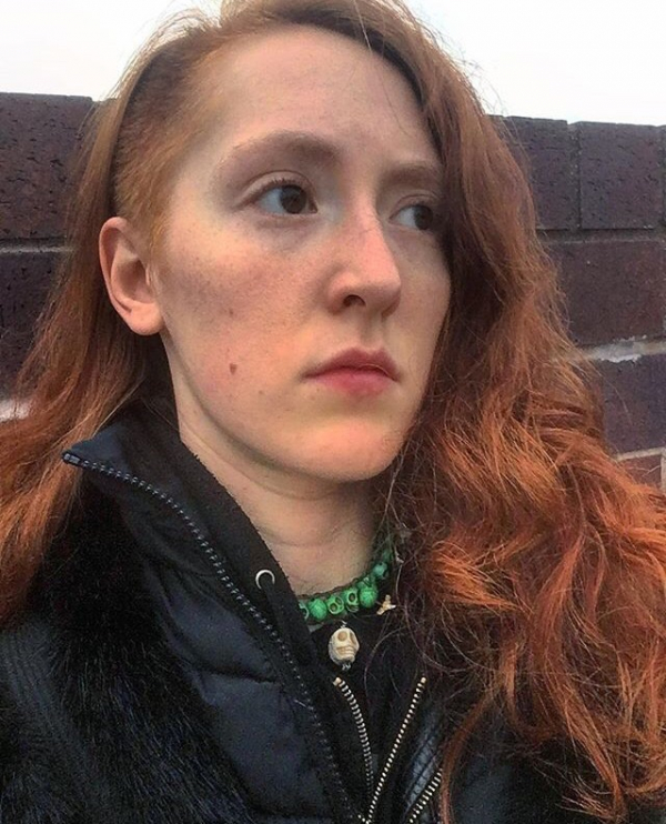 A photo of a person with long curly red hair stares off into the distance. They have the side of their head shaved, and are wearing a black coat.